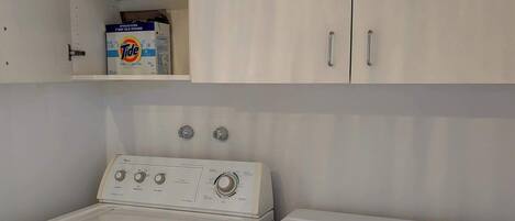A laundry area with two washing machines, a shelf with detergent bottles, and a cabinet above.