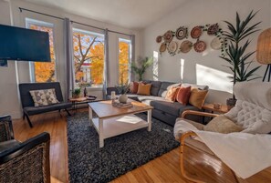 Un salon comprenant un canapé, une table basse, un téléviseur et de grandes fenêtres donnant sur l’extérieur, où l’on peut voir les feuilles d’automne.