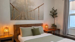 Una cama bien hecha con almohadas, una lámpara de noche, una mesita con una planta y un cuadro grande de un puente.