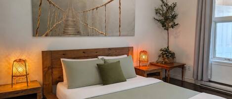 Una cama bien hecha con almohadas, una lámpara de noche, una mesita con una planta y un cuadro grande de un puente.