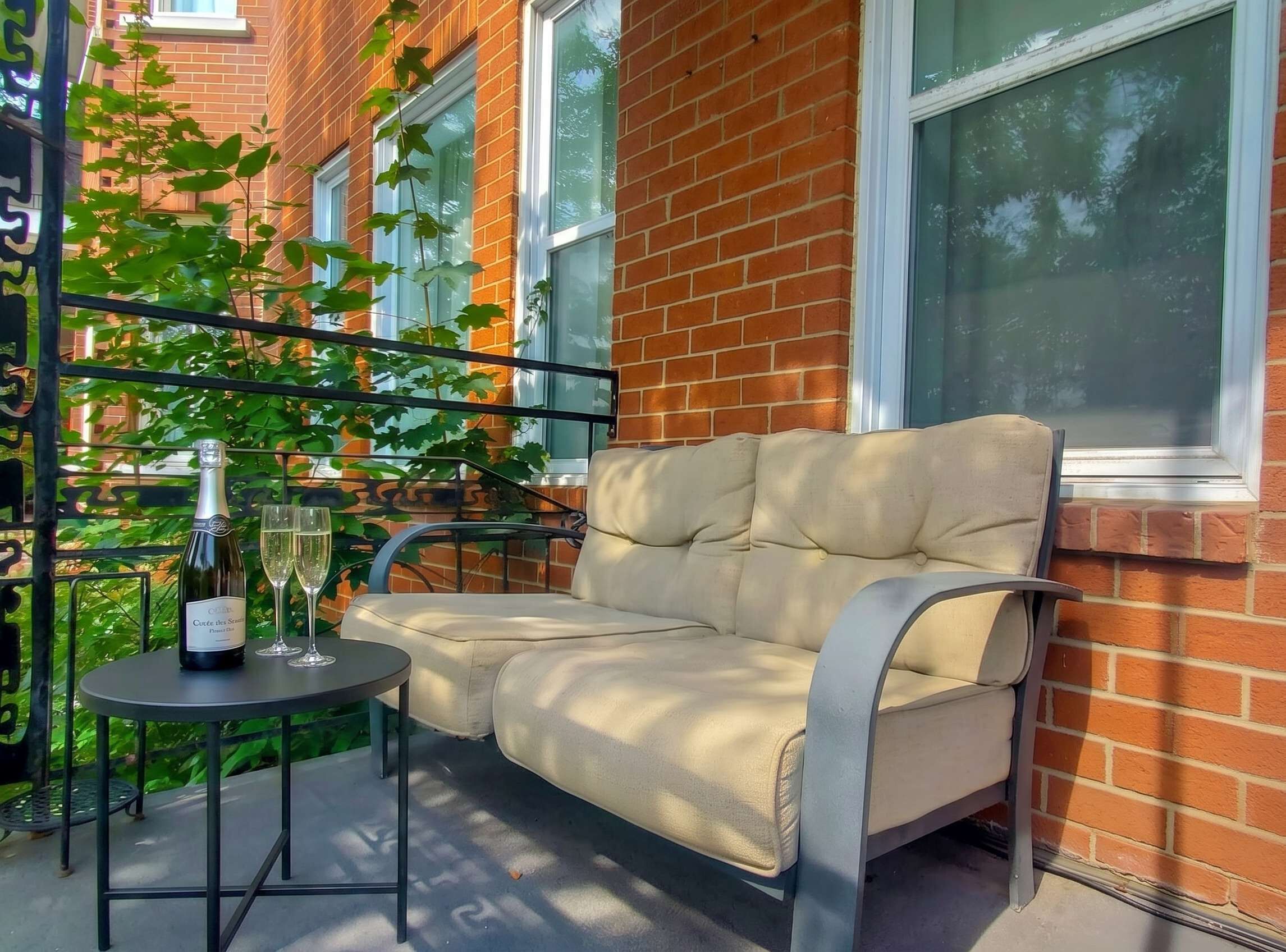 Ein Balkon mit Sitzgelegenheit mit Kissen, einem Tisch mit Flasche und Gläsern sowie Blick auf ein Backsteingebäude.