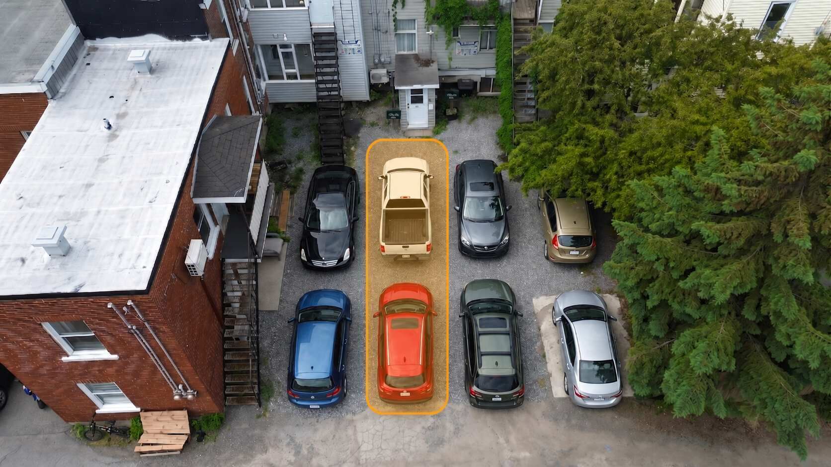 Eine Vogelperspektive auf einen Parkplatz mit verschiedenen parkenden Autos, ein Gebäude mit flachem Dach und ein Baum auf der rechten Seite.