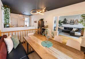 A modern living space with a wooden dining table, a black leather sofa, and a kitchen area with a refrigerator and a hanging plant.