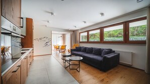 A modern kitchen with wooden cabinets, a dining area with a yellow chair, and a dark blue sofa in a bright room with large windows. - Bogdanskiego Residence Sun Snow (Zakopane)