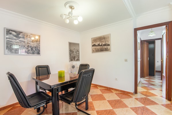 A dining area with a glass table, four chairs, a fan, and three framed pictures on the wall.