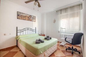 A bedroom with a bed, a desk, a chair, a ceiling fan, a window with curtains, and a wall-mounted artwork.