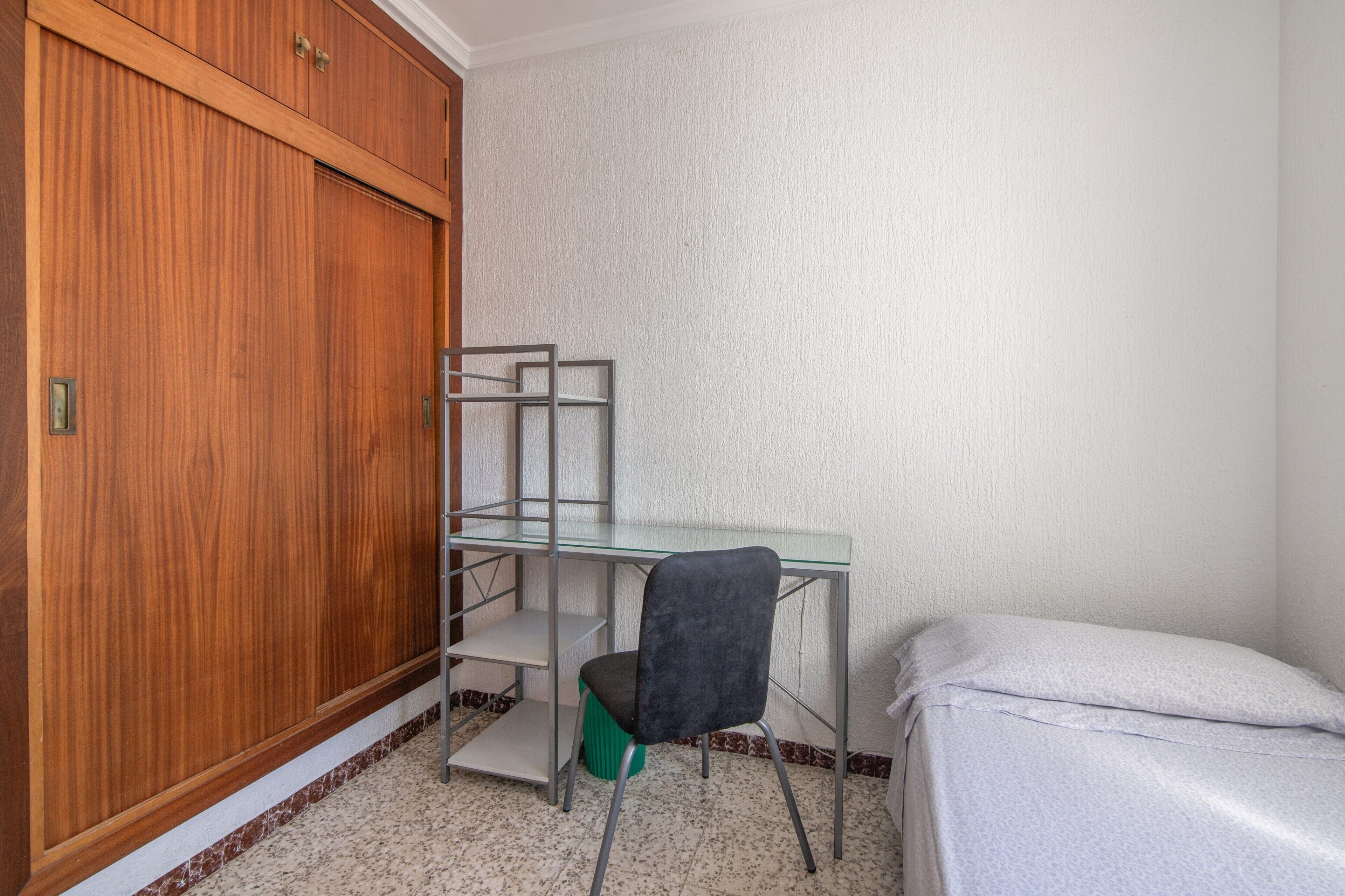 A room with a wooden wardrobe, a metal desk with shelves, a chair, and a bed with a white mattress.