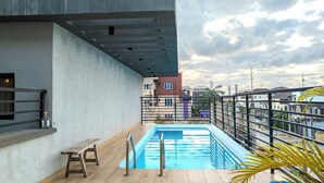 A rooftop pool with a wooden deck, metal railings, and a view of surrounding buildings. - Glassvine (Lagos)