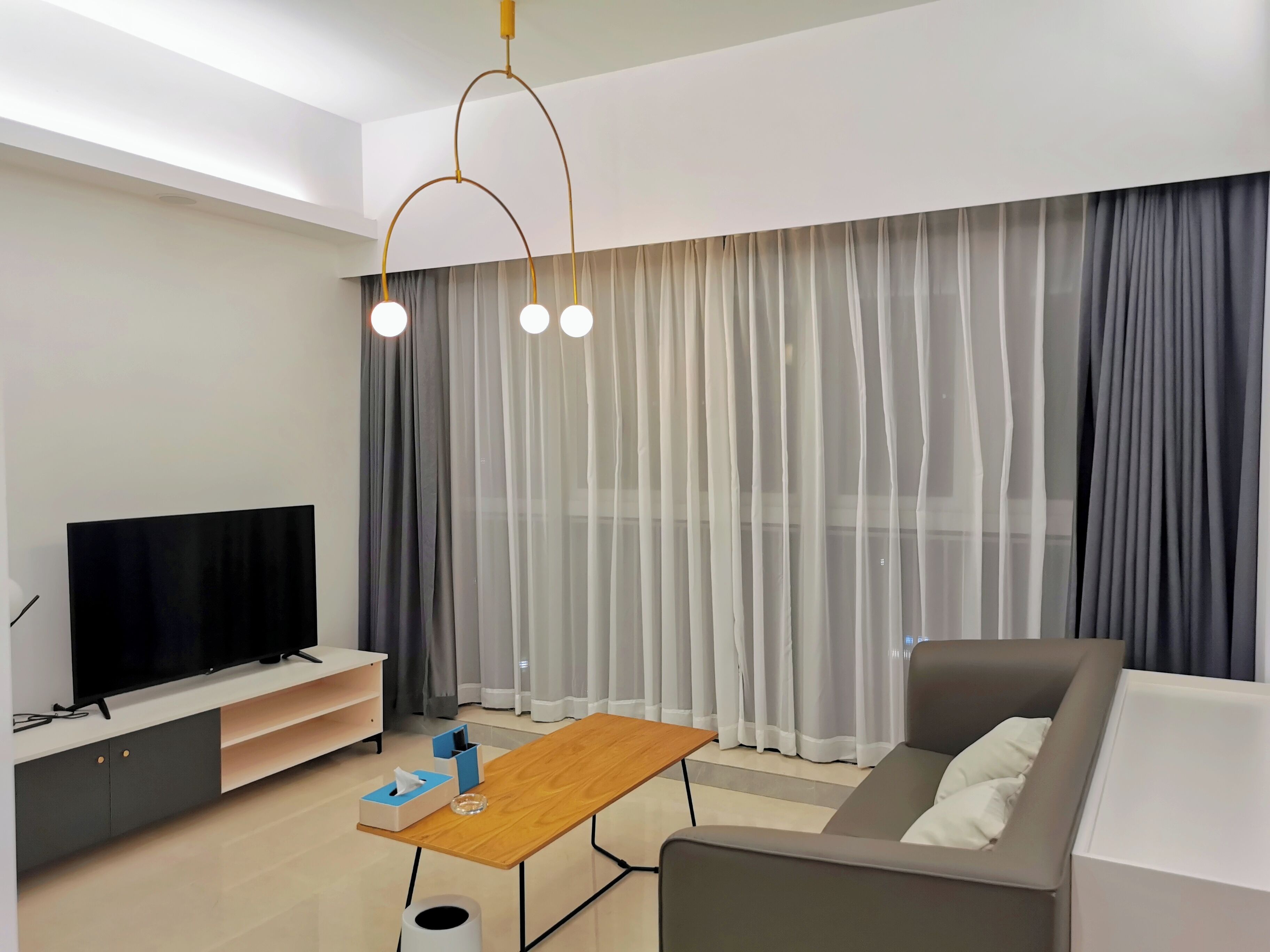 A modern living room with a flat-screen TV, a wooden coffee table, a grey sofa, and sheer curtains.