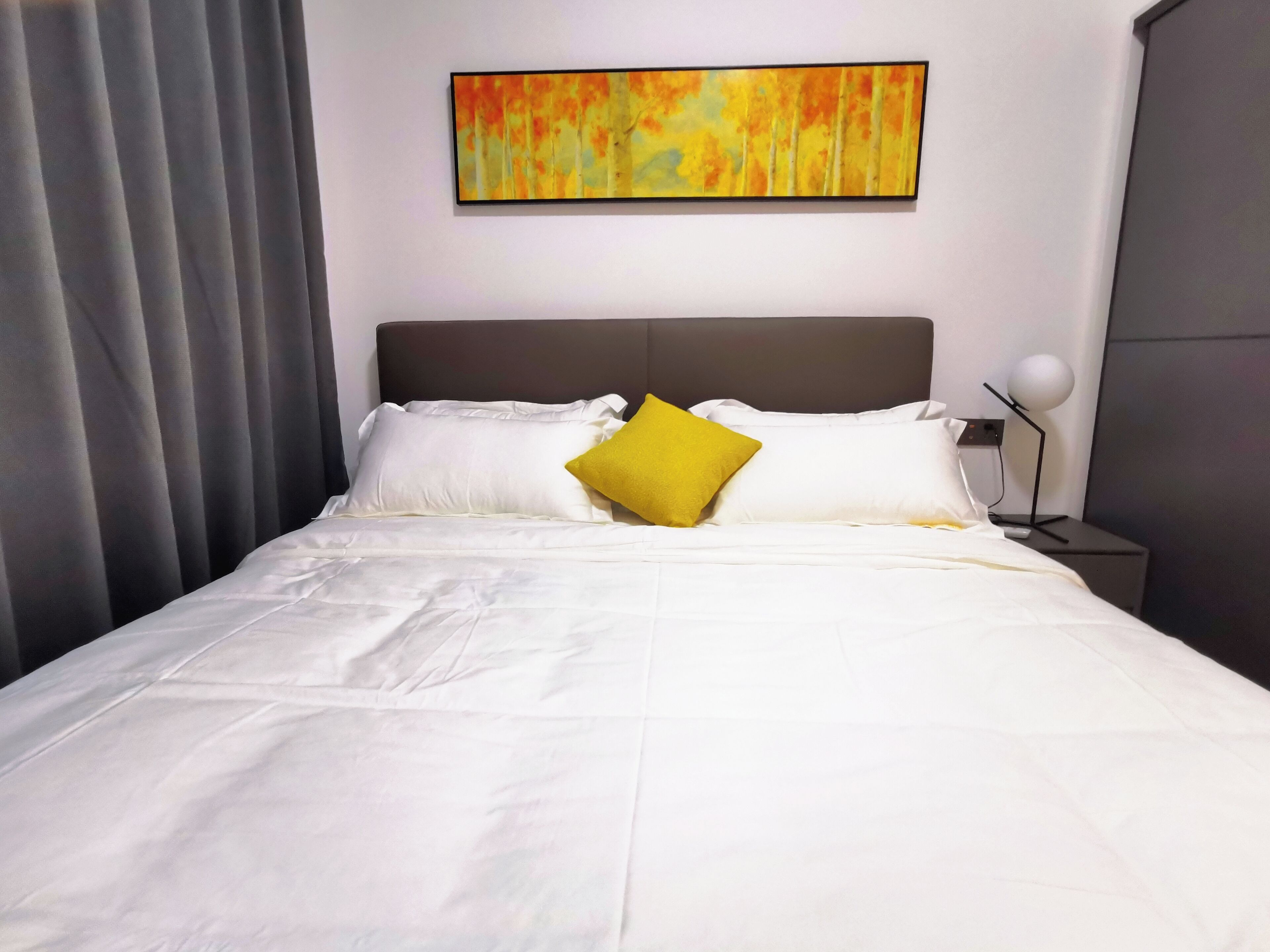 A neatly made bed with white linens and a single yellow pillow, a modern headboard, and a wall-mounted abstract painting.