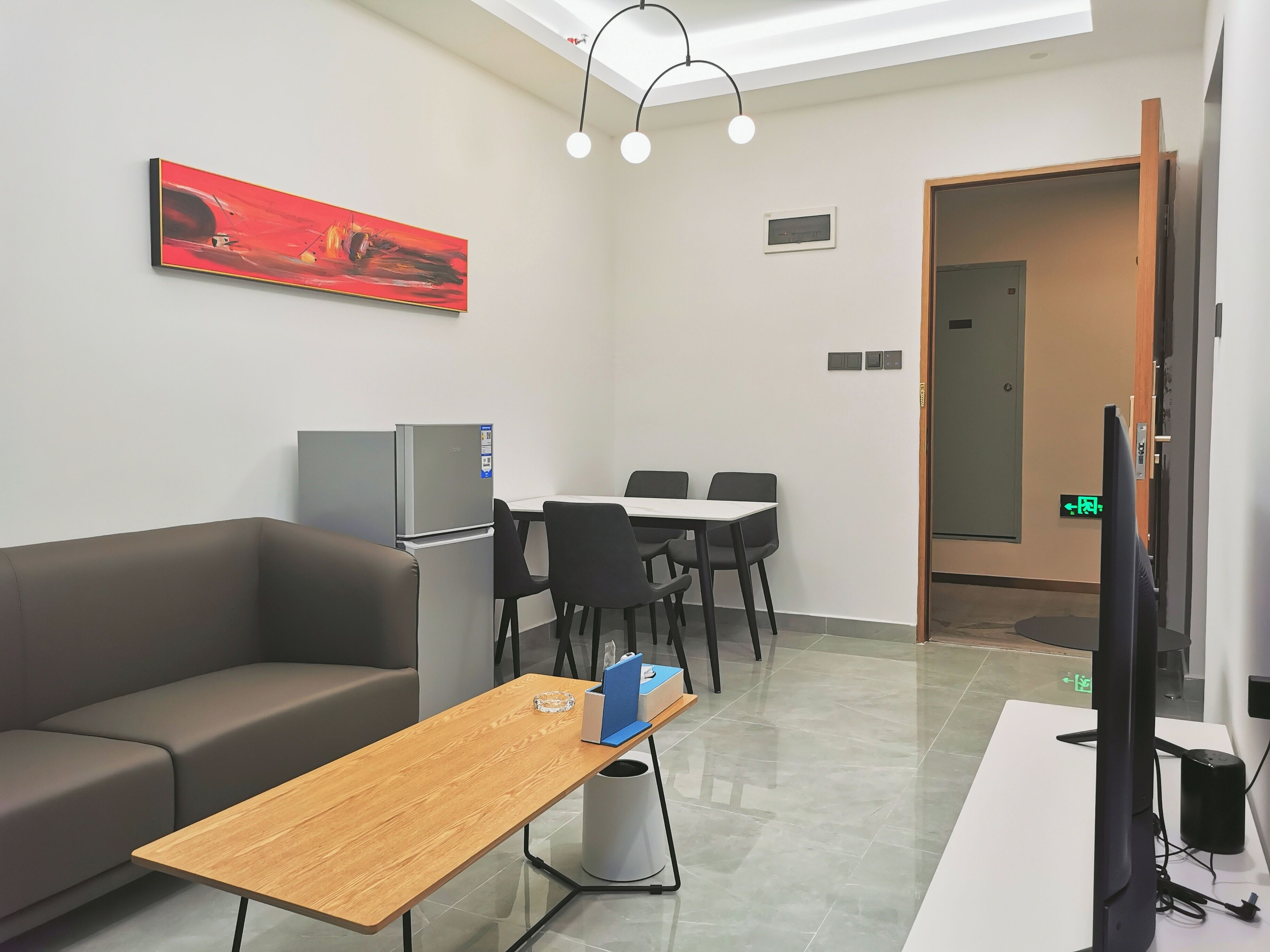 A modern hotel room with a sofa, a wooden table, a TV, a refrigerator, and a dining area with chairs and a table.