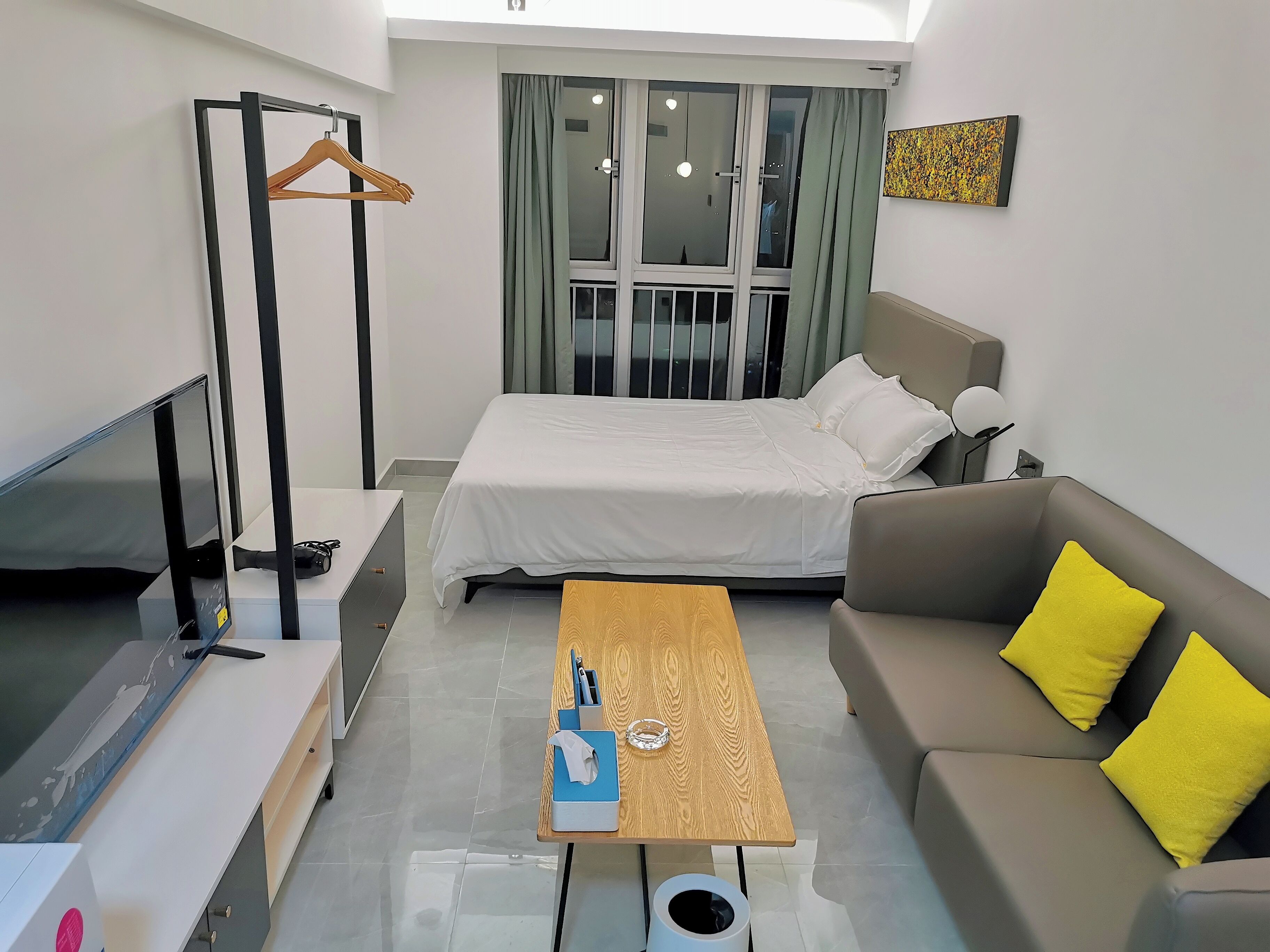 Moderni hotellihuone, jossa on sänky, sohva, puinen pöytä, televisio ja ikkuna, jossa on verhot.
