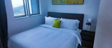 A neatly made bed with white linens and a single green pillow, a window with blinds, and a painting on the wall.