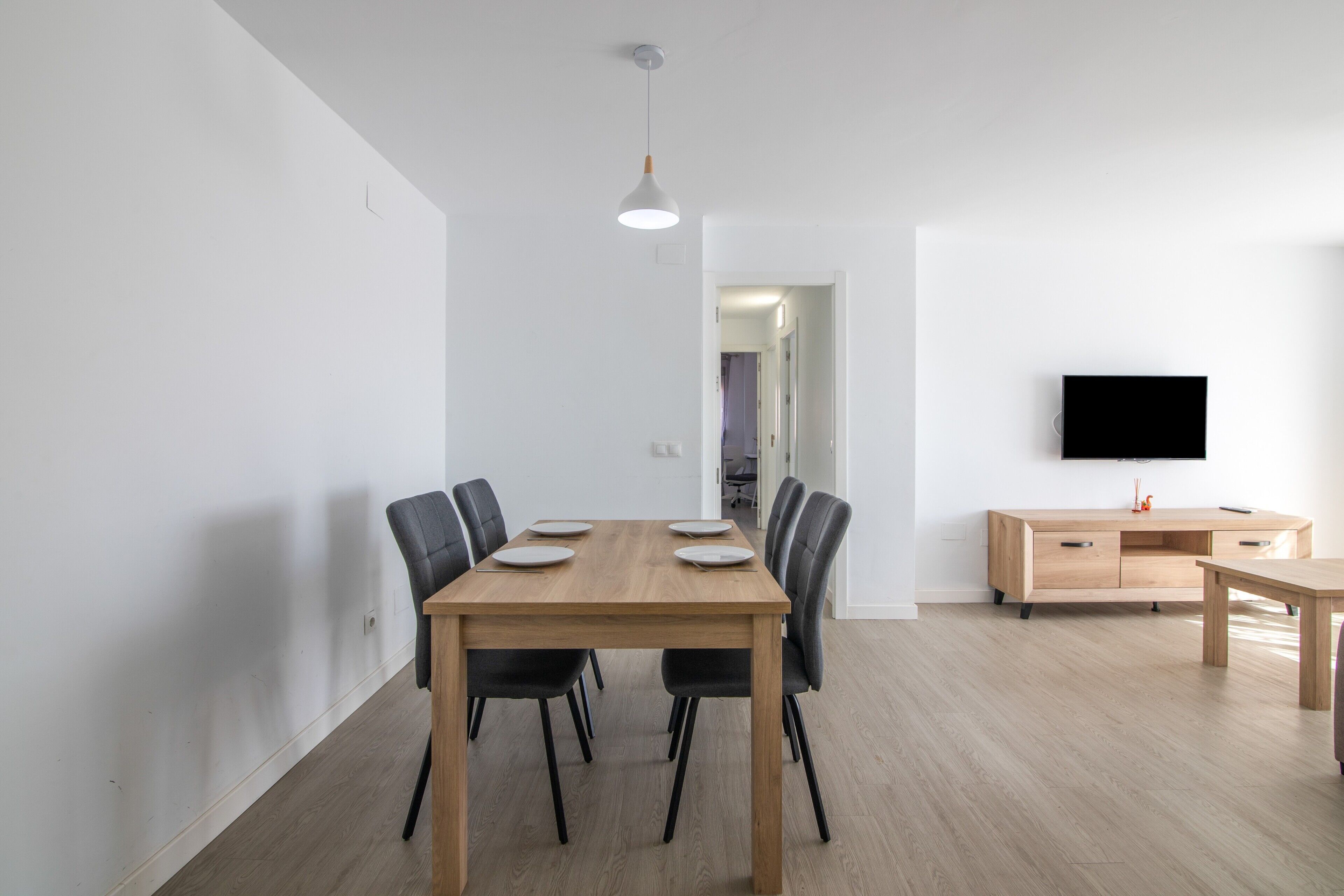 Un comedor moderno con una mesa de madera, sillas negras, una lámpara y un televisor sobre un mueble de madera, con un pasillo al fondo.