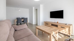 En moderne stue med sofa, sofabord i træ, fjernsyn ophængt på væggen og et spiseområde med stole og bord.