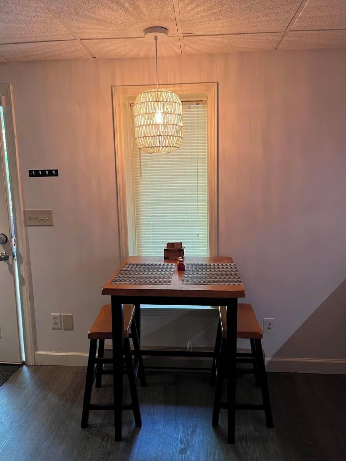 Un espace repas avec une table et des bancs en bois, un luminaire suspendu et une fenêtre avec des stores.