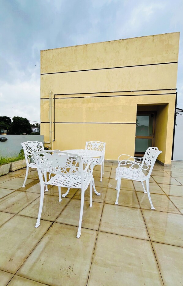 A patio with white metal chairs and a table, a yellow building in the background, and a clear sky.