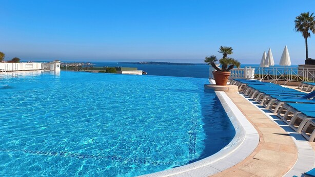 Une piscine à l’eau d’un bleu cristallin, entourée de chaises longues et de parasols, d’où l’on peut admirer l’océan et les bâtiments qui se dressent au loin.