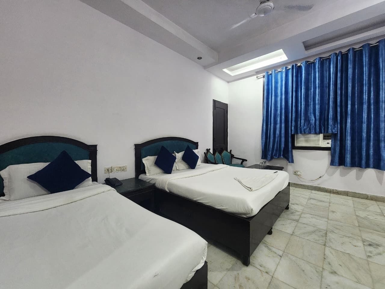 A hotel room with two beds, a ceiling fan, a window with blue curtains, and an air conditioning unit.