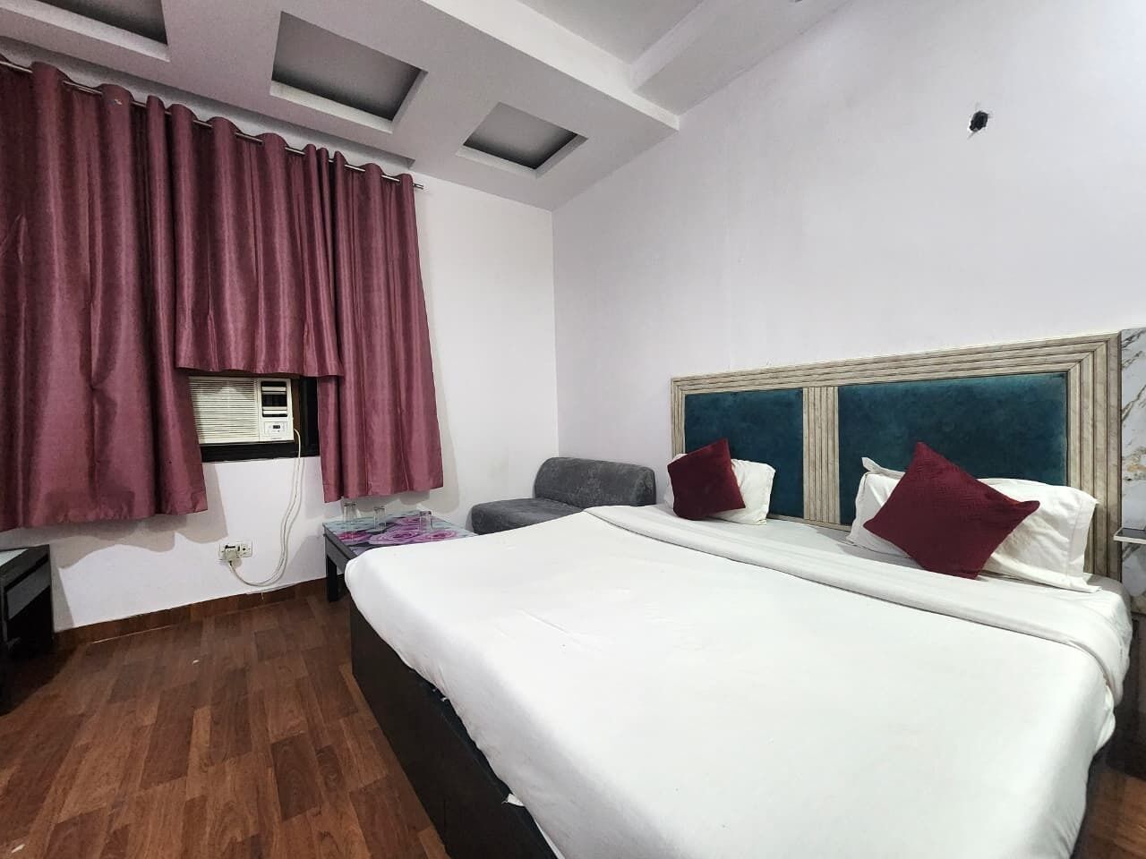 A hotel room with a large bed, a window with curtains, a small table, and a headboard with a blue accent.