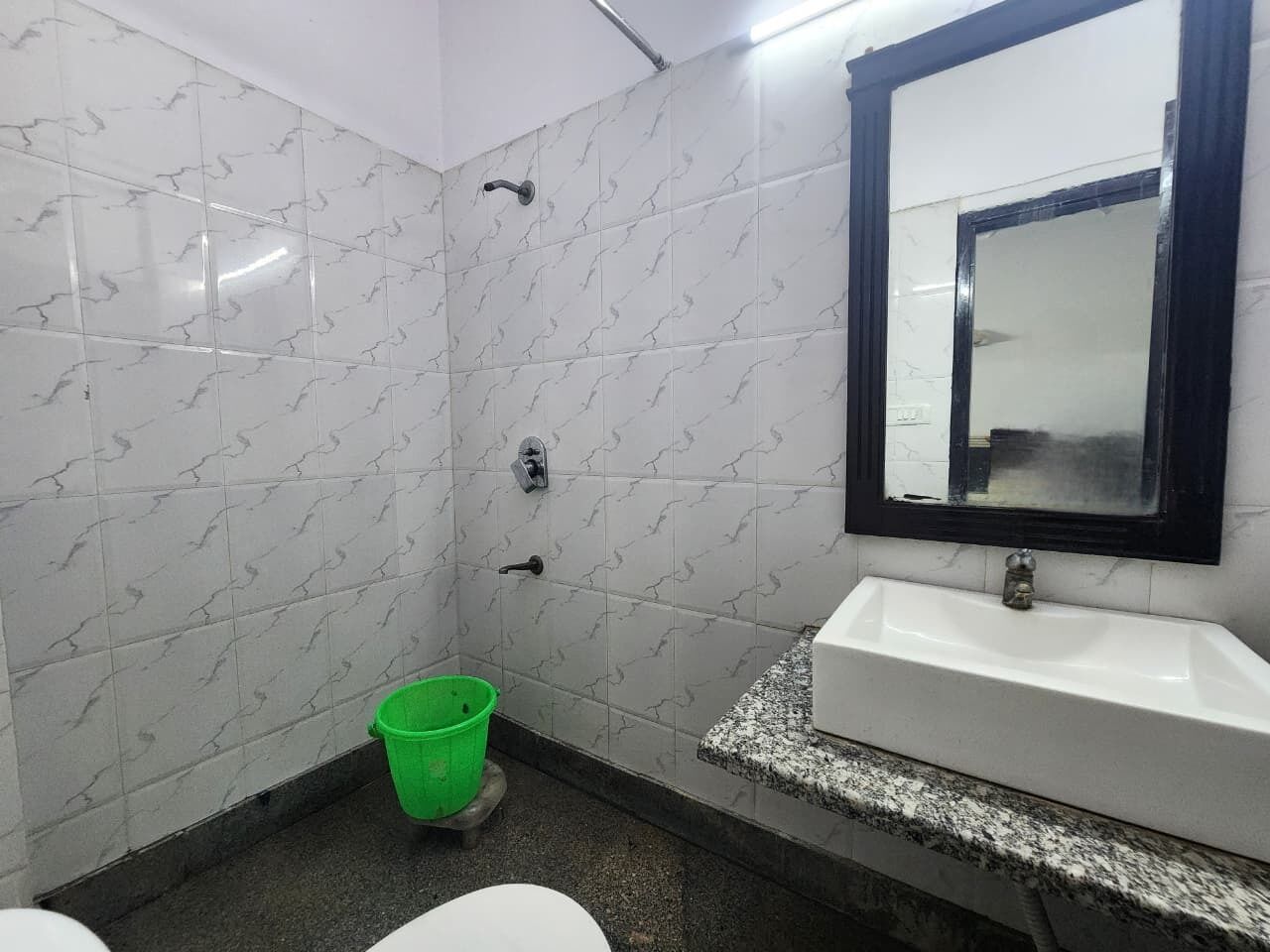 Une salle de bain avec des carreaux de marbre blanc, un seau vert, un lavabo blanc et un miroir avec un cadre noir.