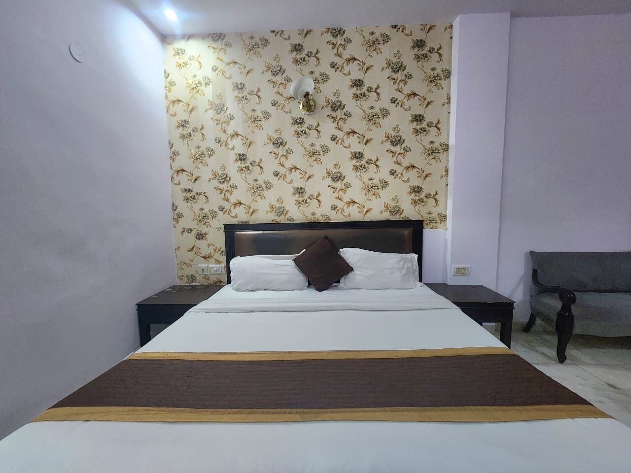 A neatly made bed with a brown throw blanket, a wooden headboard, and a floral wallpaper pattern on the wall behind.