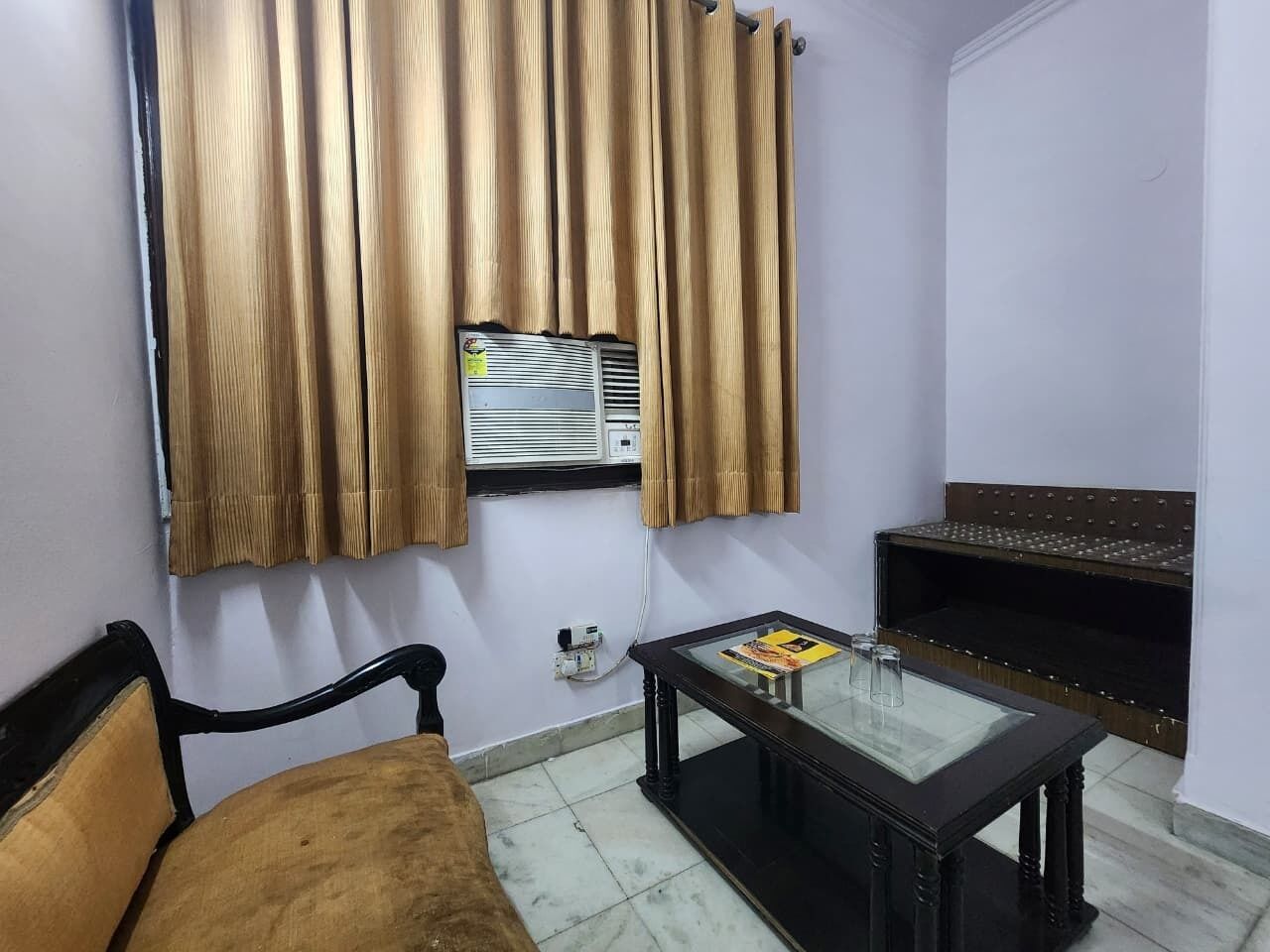 A room with a sofa, a glass-top table, a wall-mounted air conditioner, and a shelf with decorative items.