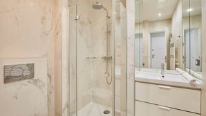 Een moderne badkamer met een douchecabine van glas, een wand betegeld met marmer en een grote spiegel boven een witte wastafel.