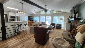A spacious living area with a brown leather sofa, a coffee table, and a pet bed. There is a kitchen with white cabinets and stainless steel appliances. The room has large windows and a ceiling fan.