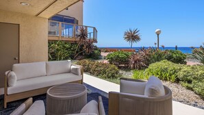 A balcony with a sofa, chairs, and a round table, overlooking a beach with plants and a clear sky.