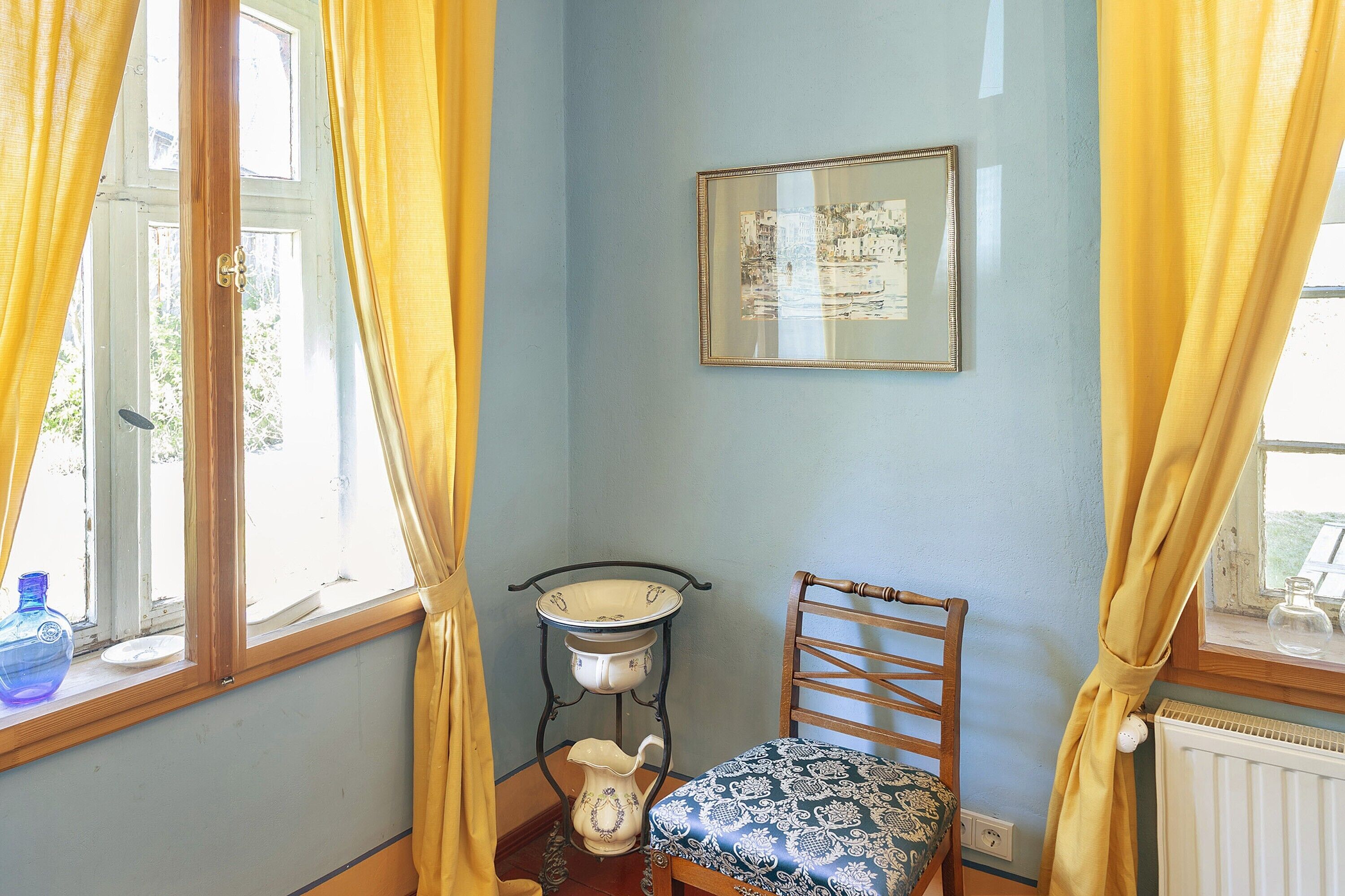 Quarto com janela, uma cadeira, mesa com jarra e quadro na parede.