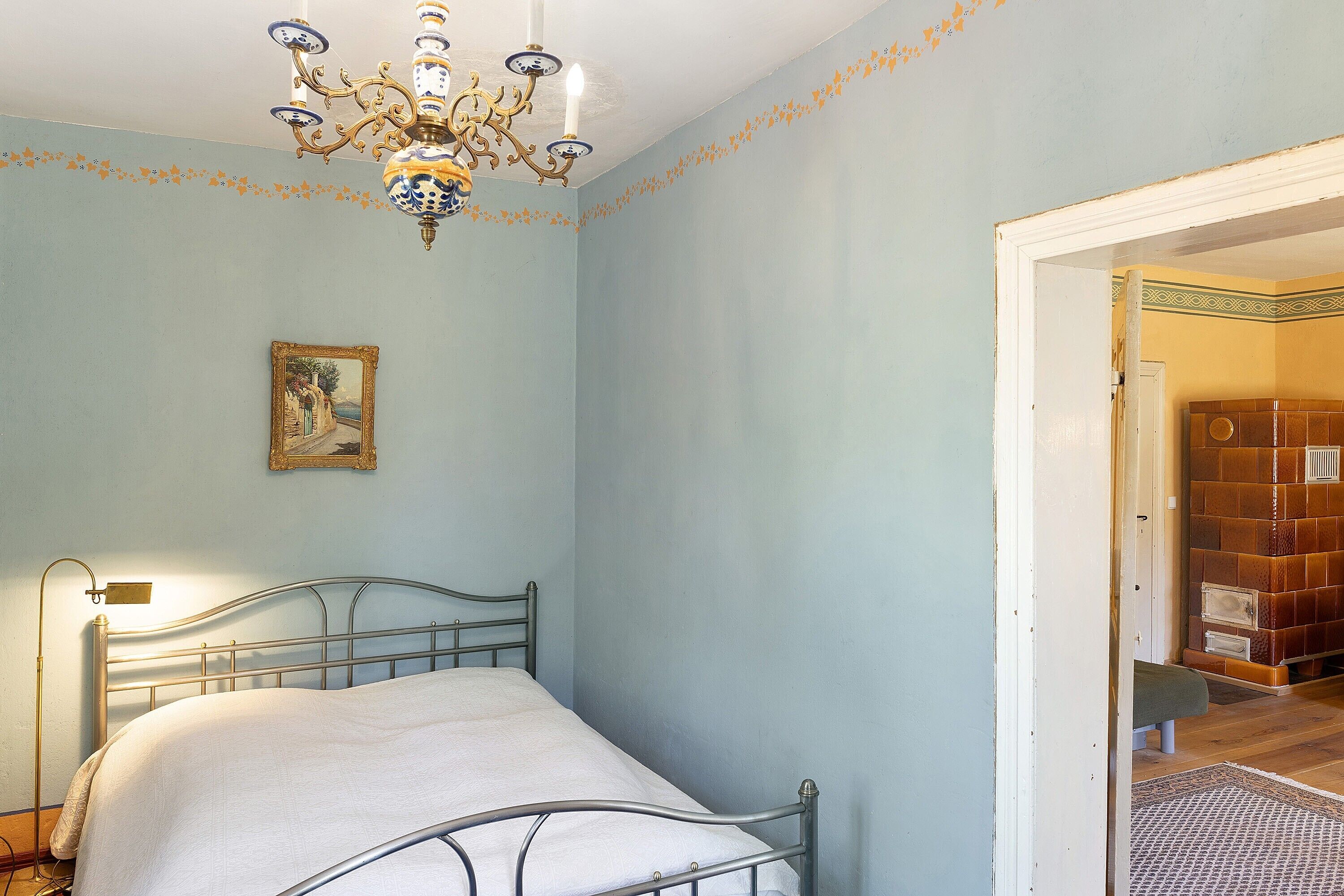 Un dormitorio con cama, candelabro, un cuadro y una puerta que da a otra habitación.