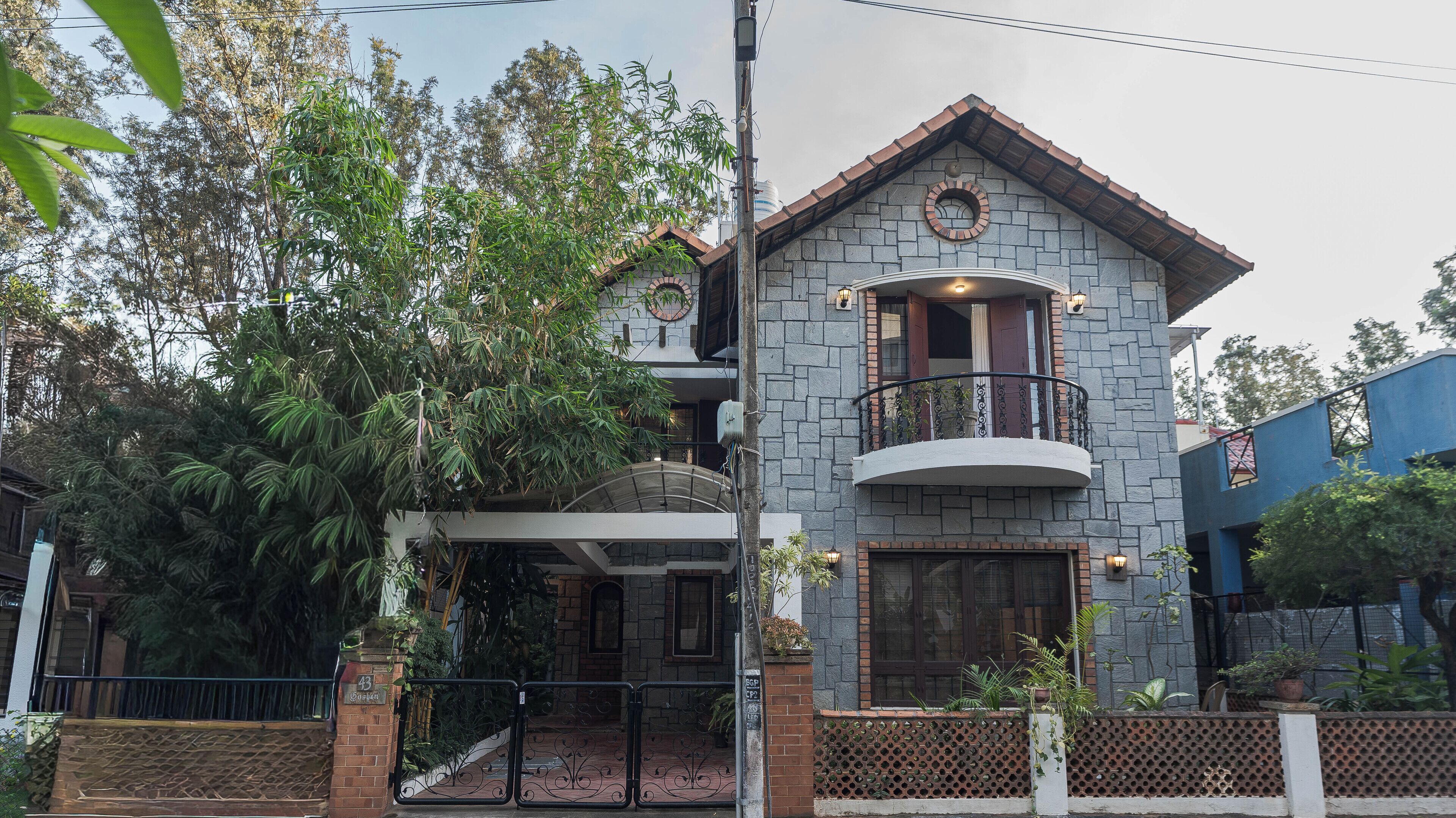Una casa de dos pisos con fachada de piedra, un balcón con barandilla de hierro forjado y un gran árbol en la parte delantera.