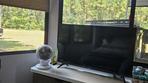 A room with a large window, a flat-screen TV on a white stand, a white fan, and a small table with bottles.