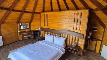 A cozy attic bedroom with a wooden sloped ceiling, a large bed, a nightstand, a chair, and a small table with a lamp.