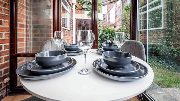 A round dining table with black plates, bowls, and wine glasses, set against a brick wall and large window overlooking a garden. - Cosy Home in Stoke on Trent (Newcastle-Under-Lyme)