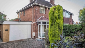 A two-story brick house with a white garage door, a conservatory, and a well-maintained garden. - Cosy Home in Stoke on Trent (Newcastle-Under-Lyme)