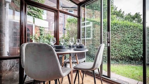 A dining area with a table set for four, surrounded by large windows offering a view of a garden and neighboring buildings. - Cosy Home in Stoke on Trent (Newcastle-Under-Lyme)