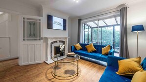 A modern living room with a blue sofa, a glass coffee table, a fireplace, and a large window with curtains. - Cosy Home in Stoke on Trent (Newcastle-Under-Lyme)