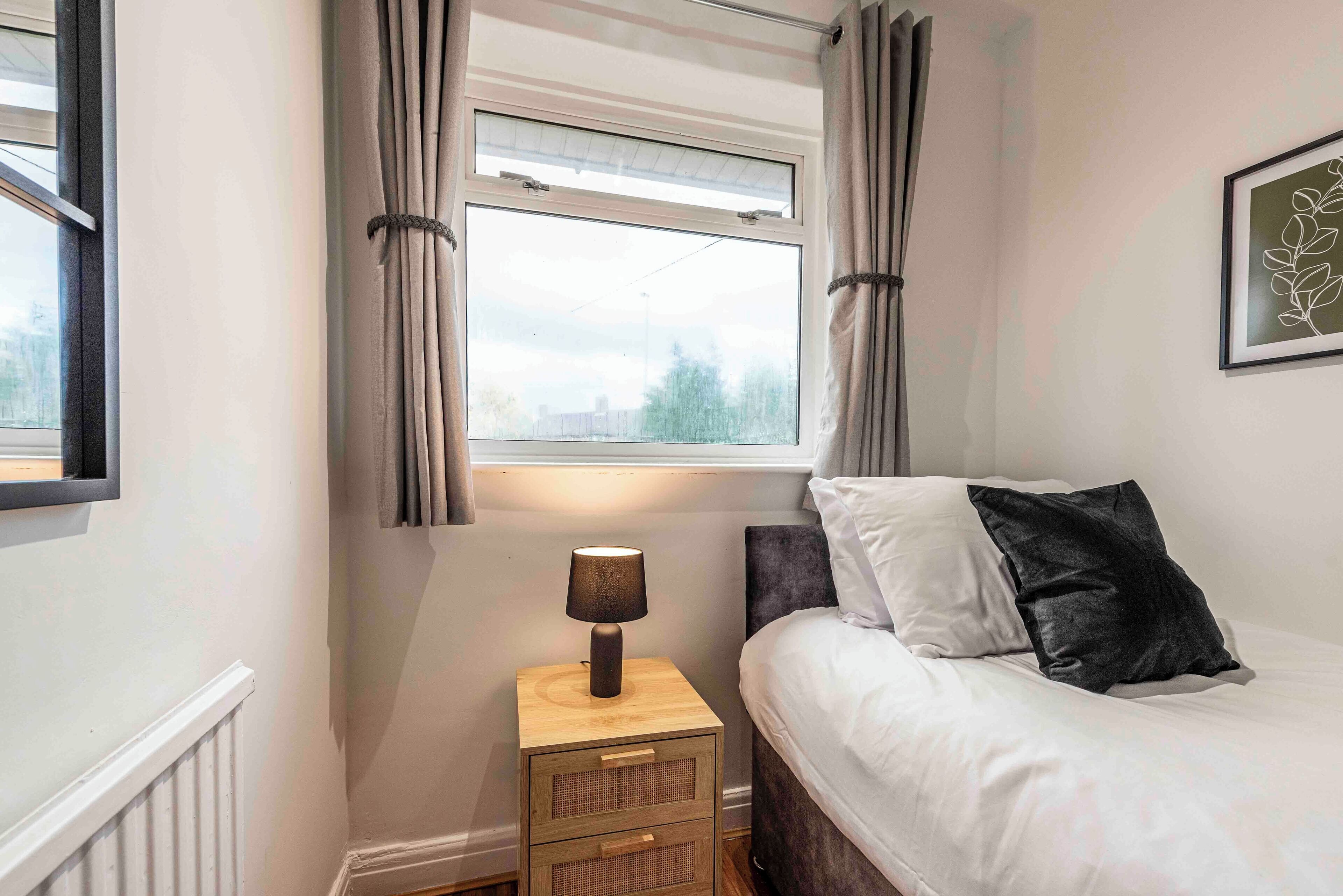 A bedroom with a bed, a bedside table with a lamp, a window with curtains, and a framed picture on the wall.