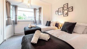 A hotel room with two beds, a window with curtains, and framed pictures on the wall. - Cosy Home in Stoke on Trent (Newcastle-Under-Lyme)