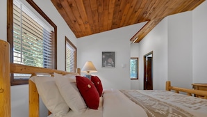 A bedroom with a wooden ceiling, a bed with white and red pillows, a lamp, and a patterned throw. - Snowflake Chalet -Govy Chalet Steps to Glade Trail (Government Camp)
