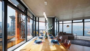 Un comedor moderno con una larga mesa de madera dispuesta para una comida, ventanas amplias que ofrecen vistas a la ciudad y una zona de estar acogedora con sofá y sillones.