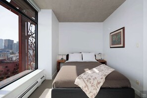 A modern bedroom with a large window, a bed with striped bedding, a nightstand with a lamp, and a framed picture on the wall. - Elegant 2-Bedroom Condo in Vancouver (Vancouver)