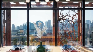 Una mesa de comedor con platos, vasos y una botella, con vistas a un paisaje urbano a través de amplios ventanales.