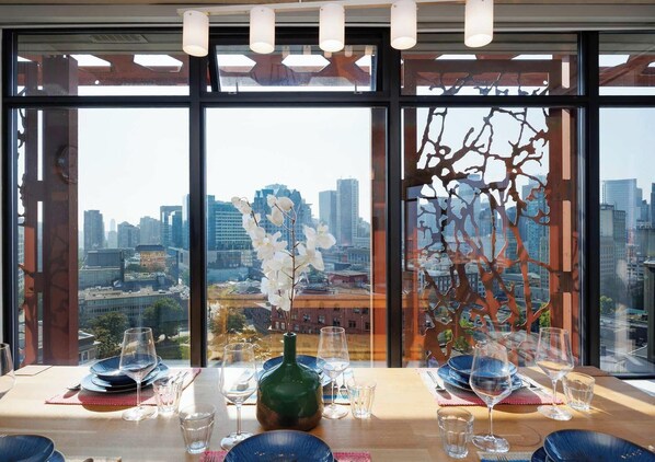 Una mesa de comedor con platos, vasos y una botella, con vistas a un paisaje urbano a través de amplios ventanales.