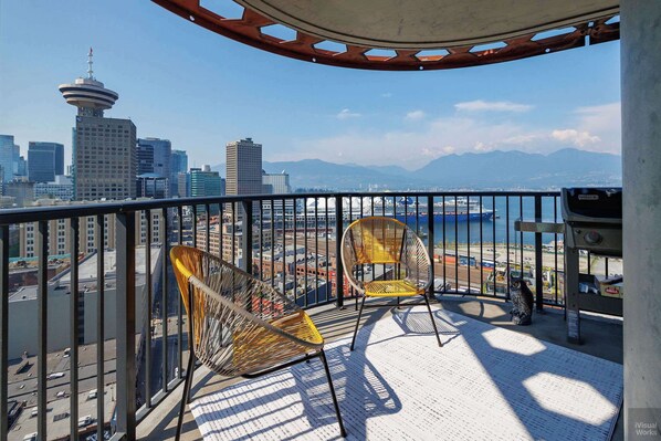 A balcony with two wicker chairs, a view of a cityscape including a tower, and a body of water with a ship. - Elegant 2-Bedroom Condo in Vancouver (Vancouver)