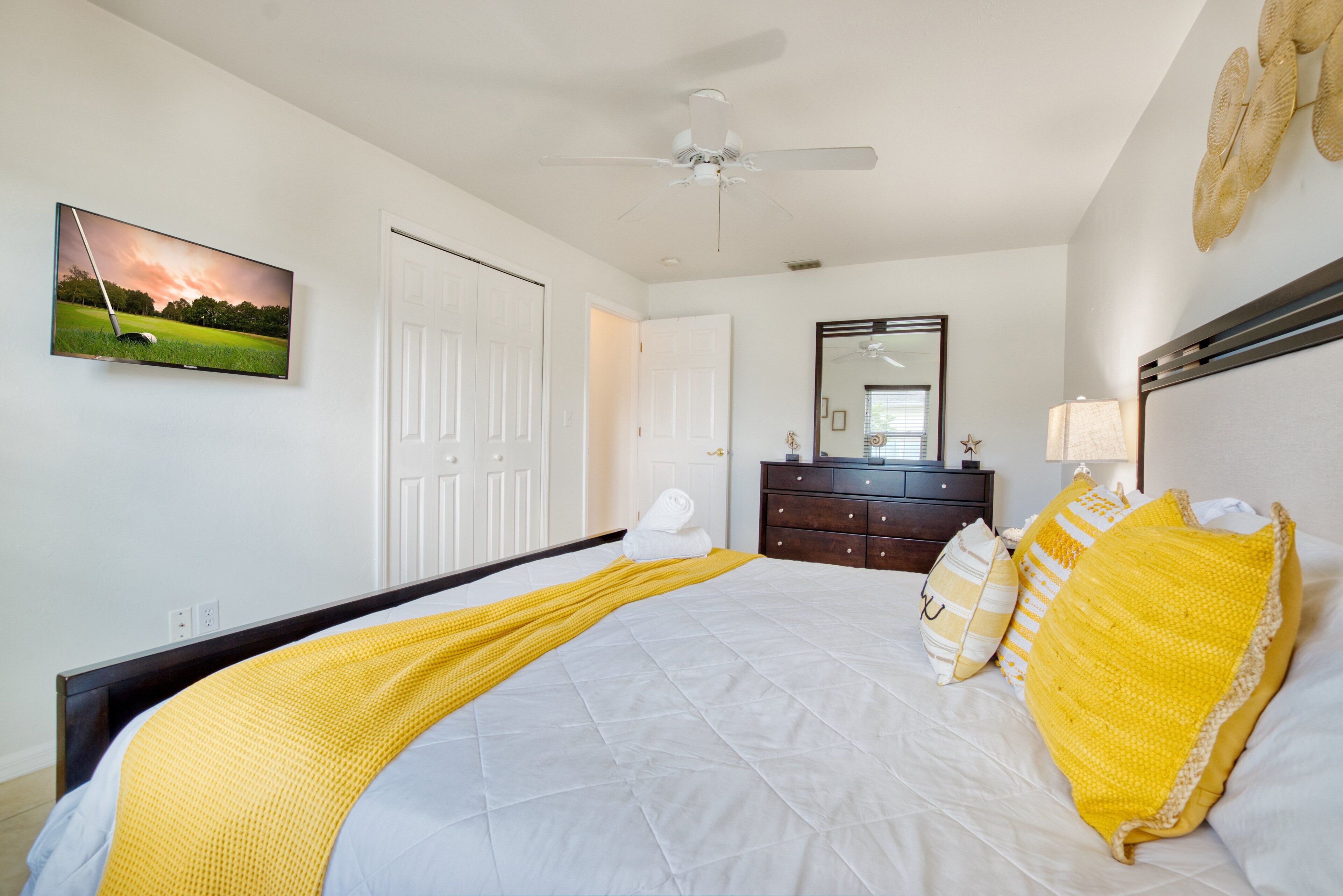 A bedroom with a bed, a ceiling fan, a dresser, a mirror, and a television on the wall.