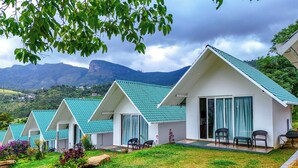 A row of white cottages with green roofs, each with a patio and seating area, set against a mountainous backdrop. - Gardenia Kanthalloor by VOYE HOMES (Devikolam)