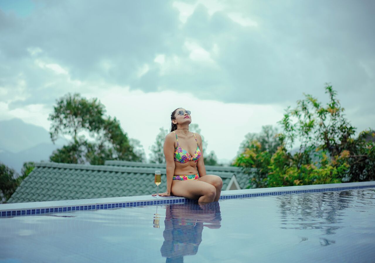 A person sitting on the edge of a swimming pool with a drink in hand, surrounded by greenery and a cloudy sky.